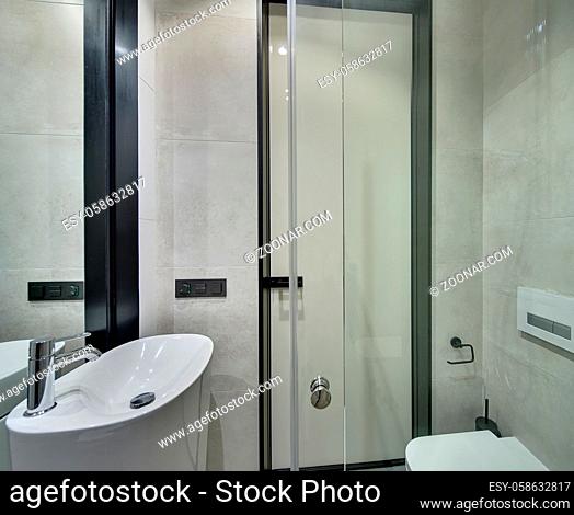 Modern style bathroom with light tiles on walls. There is a white sink with chrome faucet, glass door, mirror, toilet, entrance door. Horizontal