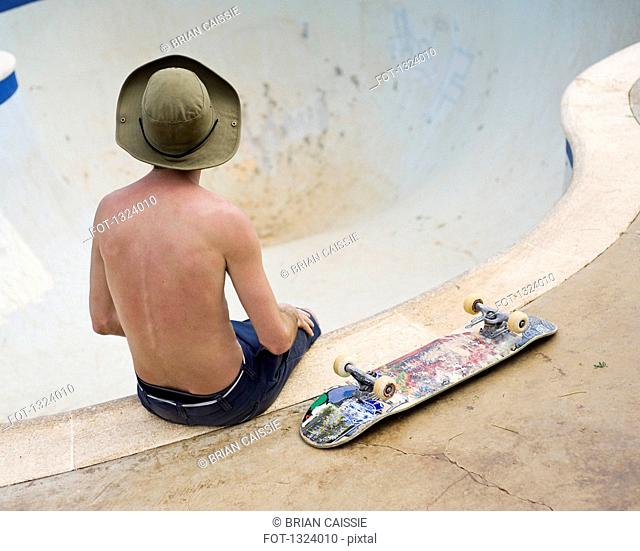 Rear view of man with skateboard sitting on the edge of a skateboarding bowl