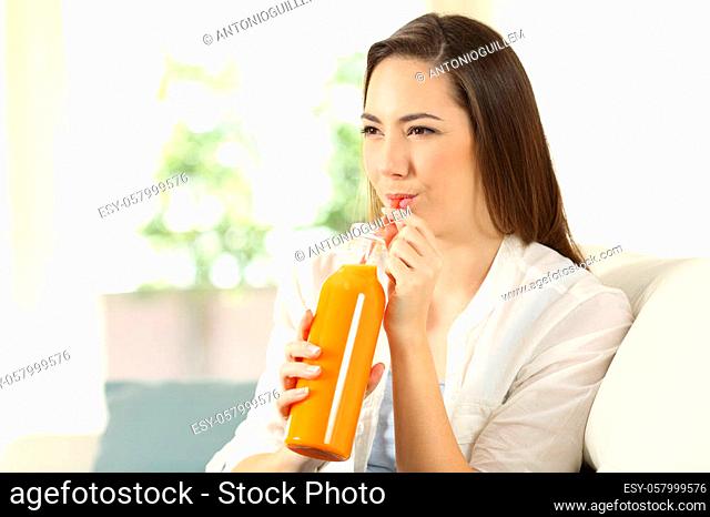Woman drinking orange juice with a straw sitting on a couch in the living room at home