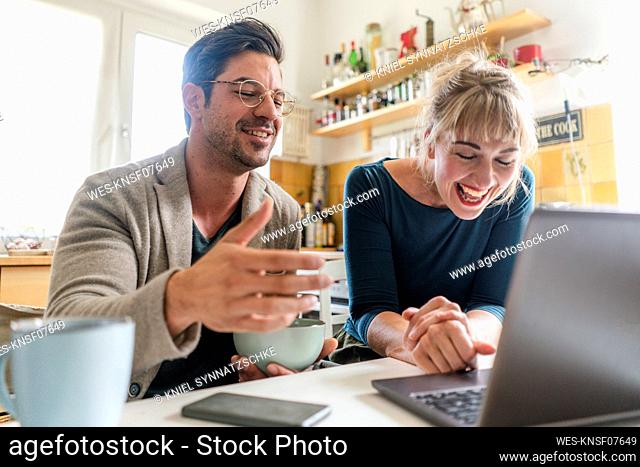 Happy couple sitting at table in kitchen using laptop