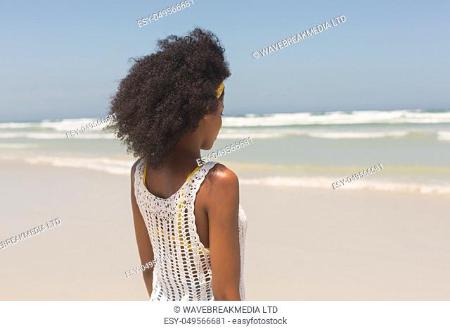Rear view of young African American woman standing on the beach in the sunshine. She is looking away