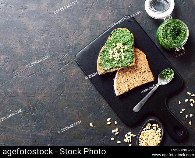 whole grain rye bread with fresh basil pesto sauce on black wooden cutting board over dark concrete background. Top view or flat-lay. Copy space