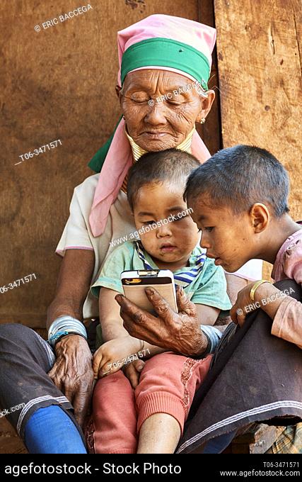 Kayan Lahwi woman with brass neck coils and traditional clothing looking at cellphone with her grandchildren. The Long Neck Kayan (also called Padaung in...