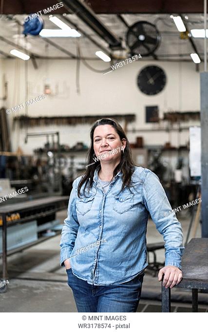 Woman with brown hair wearing Denim shirt standing in metal workshop, smiling at camera