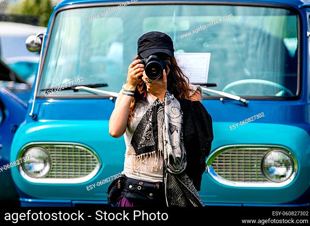 selective focus on young caucasian photographer woman holds DSLR camera in front of old blue van, wearing black hat scarf, during a antique cars show