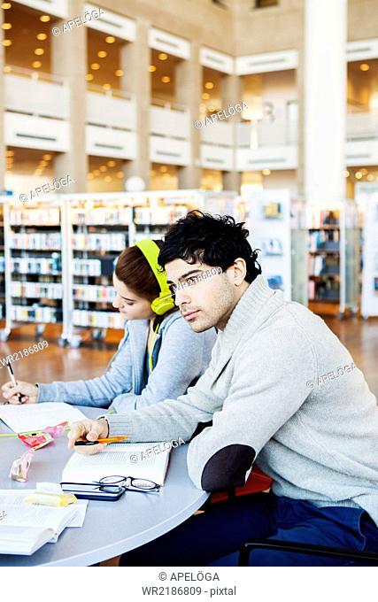 Thoughtful young man looking away while friend reading notes at table in library