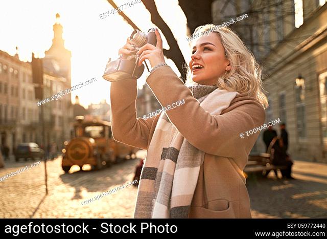 Young beautiful cheerful blond woman wearing scarf and beige coat taking photos with her vintage film camera on a sunny day at Rynok square in Lviv, Ukraine