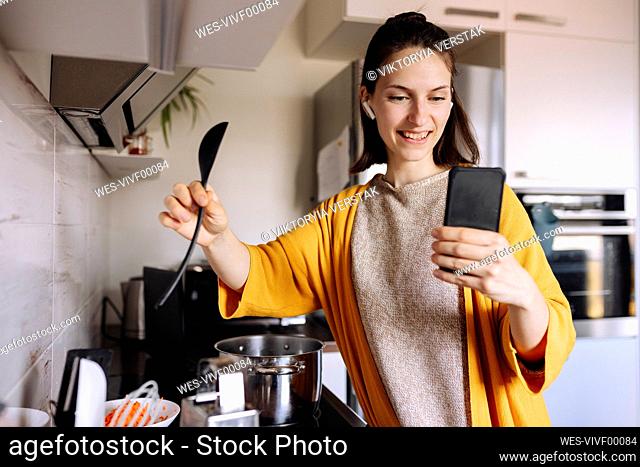Smiling young woman holding wooden spoon having video call through mobile phone in kitchen