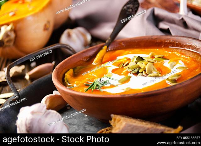 Clay dish with homemade rustic pumpkin soup with seeds on wooden table with bread and greens aside