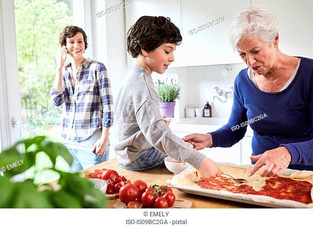 Grandmother and grandson making pizza in kitchen, mother in background using smartphone