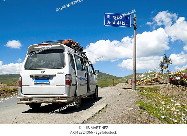 A tourist van on the road with prayer flags on the way to Litang, a Tibetan village in the west of Sichuan province, passing mountains with high altitude;...