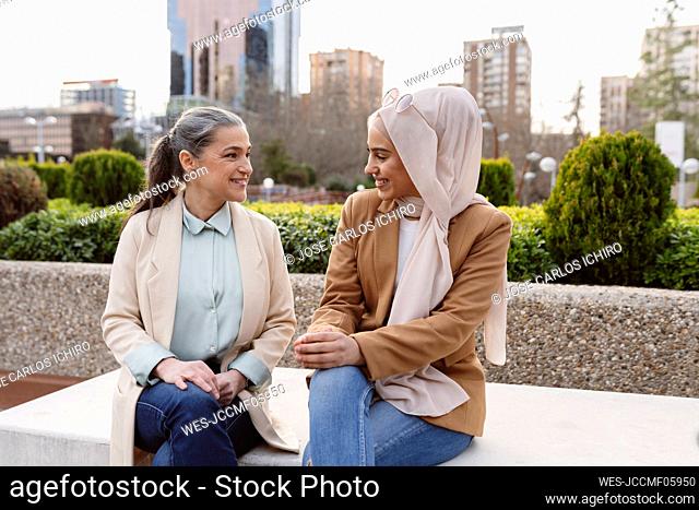 Smiling businesswomen talking with each other sitting on bench at office park