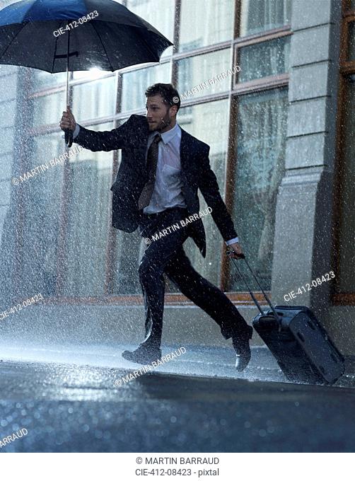 Businessman with suitcase and umbrella crossing rainy street