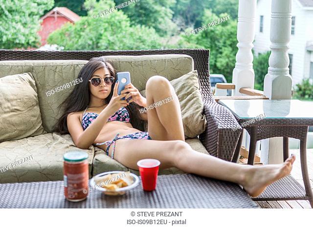 Teenage girl wearing bikini reclining on patio sofa reading smartphone