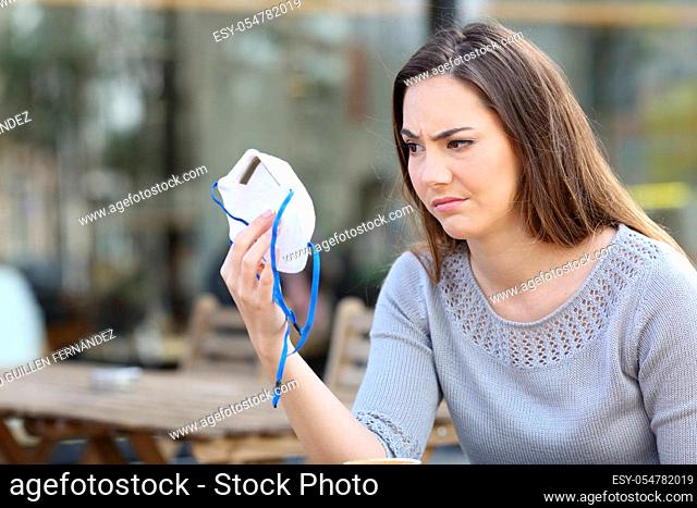 Doubtful woman looking hesitant at protective mask sitting on a coffee shop terrace