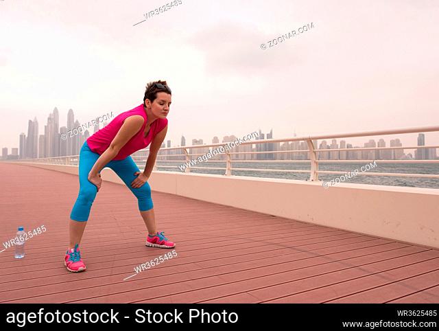 very active young beautiful woman stretching and warming up on the promenade along the ocean side with a big modern city in the background to keep up her...