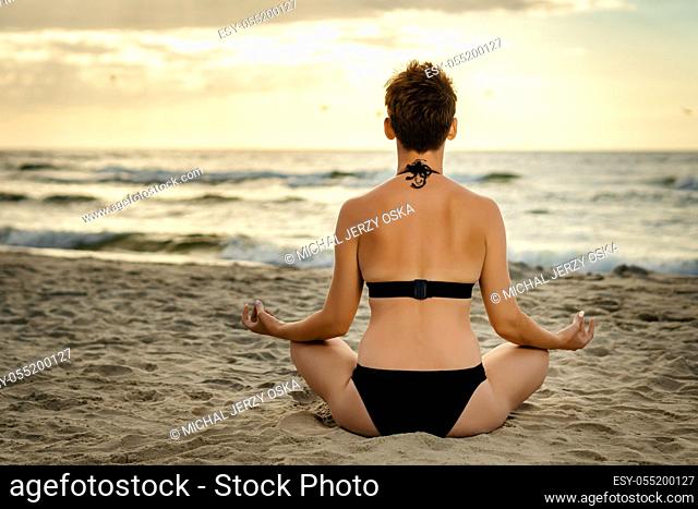beautiful woman in a black swimsuit meditating on the beach at sunset