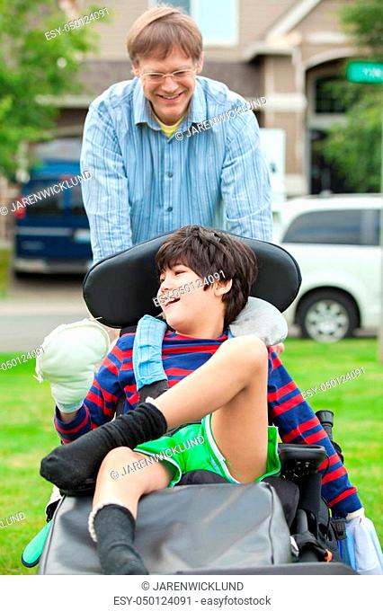 Caucasian father pushing disabled biracial ten year old son in wheelchair outdoors. Child has cerebral palsy