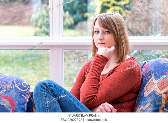Young woman dressed in brown sweater is sitting on the couch and pensively looking at the camera
