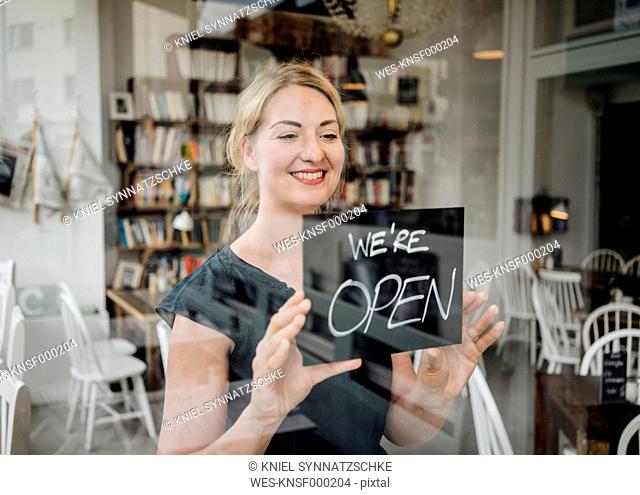 Smiling woman in a cafe attaching open sign to glass pane