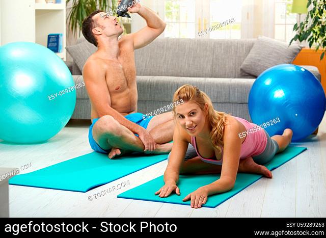 Young women doing streching exercise on fitness mat, muscular man drining water in the background. Selective focus on woman
