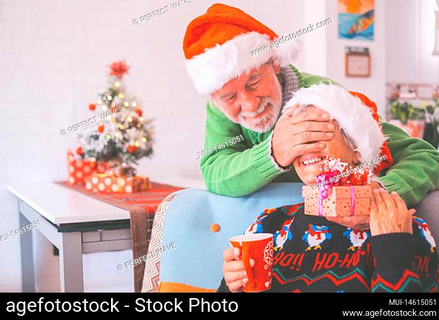 Senior couple in warm clothing and santa hat, man covering eyes of old wife with a surprise gift for her. Loving old romantic heterosexual couple celebrating...