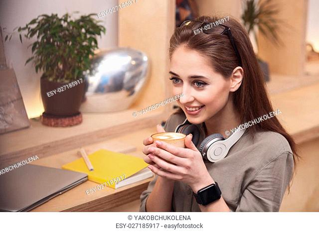 Attractive girl is enjoying a cup of latte. She is sitting and smiling happily