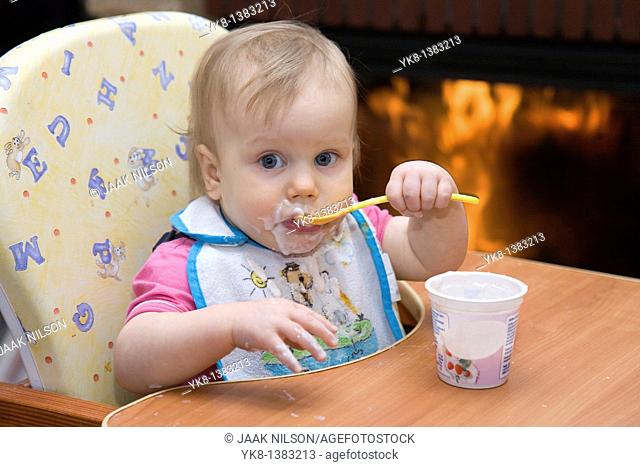 One year old dirty baby girl in high chair eating yogurt using spoon