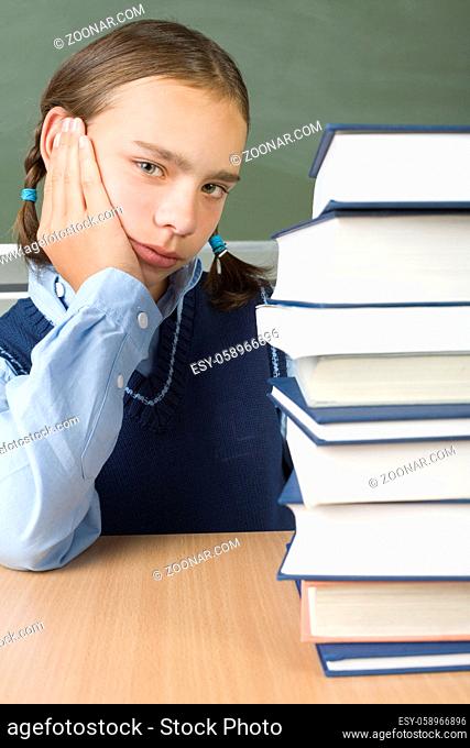 Young, sad girl sitting at desk nearby books. Looking at camera, front view