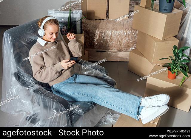 Woman with headphones using mobile phone on plastic wrapped armchair