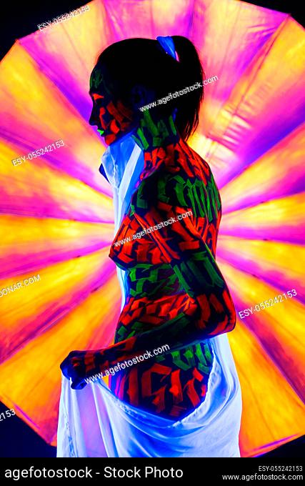 Woman with artistic UV bodyart with white ribbon in her hair and white cloth around her waist full-length reraview against colorful background