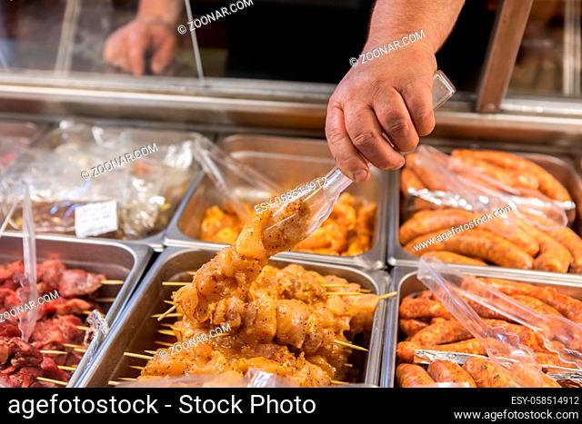 Butcher hand picking a wooden skewer of poultry meat ready for cooking using a plastic tong in close up view. bistro restaurant display refrigerator