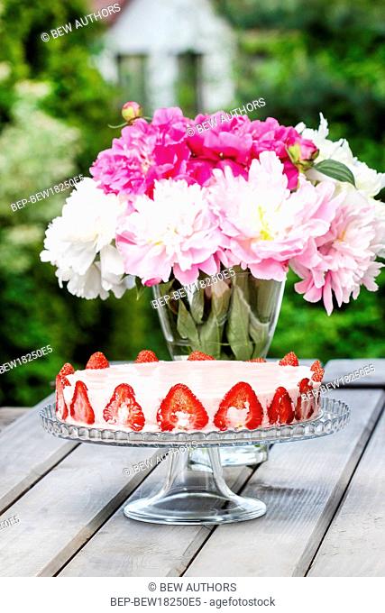 Strawberry cake on rustic wooden table in lush summer garden. Pink peonies in the vase. Selective focus