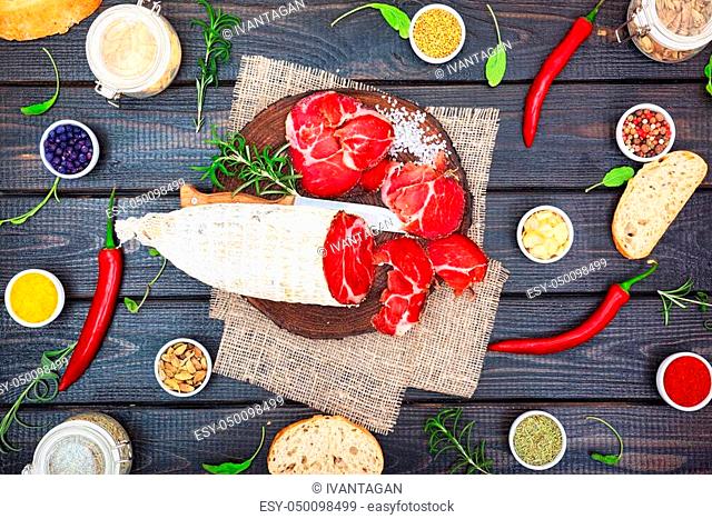 Sliced cured coppa with spices and a sprig of rosemary on dark wooden rustic background
