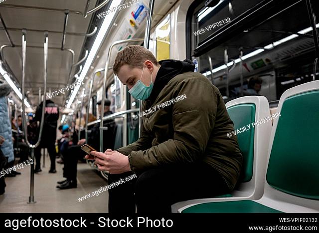 Young man in commuter train, wearing face mask, using smartphone