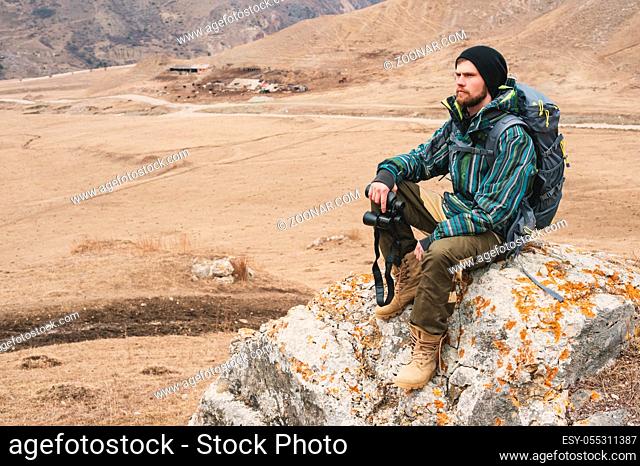 Tired bearded hipster with binoculars in his hands sits on a stone among the Caucasian mountains and looks out into the distance