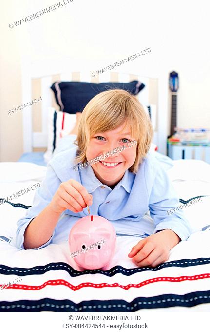 Smiling boy inserting a coin in a piggybank lying down on bed