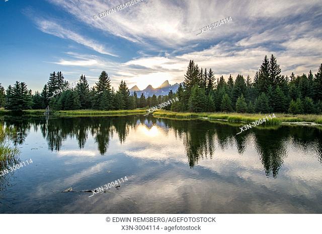 Sunlight creeps behind peaks of Teton Mountain Range with glassy water in the foreground, Grand Tetons National Park, Teton County, Wyoming. USA