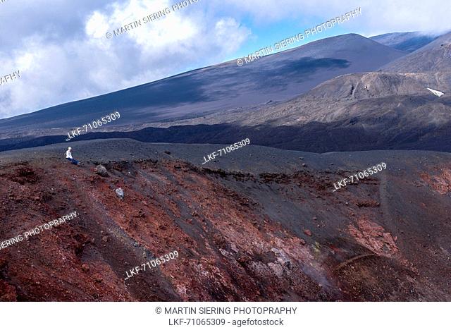 Woman sitting at the edge of an extinct crater at the foot of the Etna volcano. Minerals in the soil are the reason for the red color of the rocks, Sicily