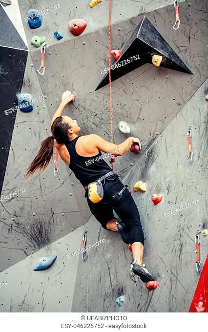 Beautiful young woman in black outfit climbing on practical wall in gym, bouldering, extreme sport, rock-climbing concept