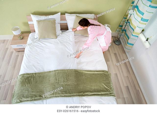 Young Female Housekeeper Arranging Bedsheet On Bed In Room