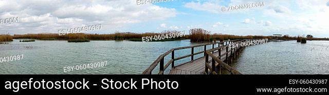 A panorama of a long wooden pier and boardwalk in brackish water wetlands with esparto grass and lagoon under an overcast sky