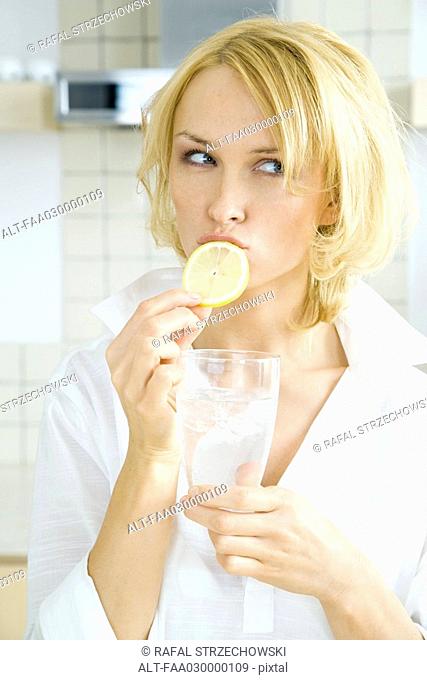 Young woman biting into a slice of lemon, looking away, holding a glass of water