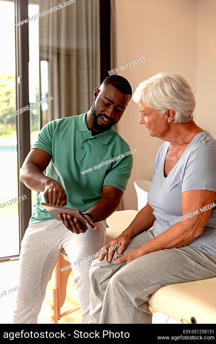 African american male physiotherapist showing digital tablet to caucasian senior woman at home