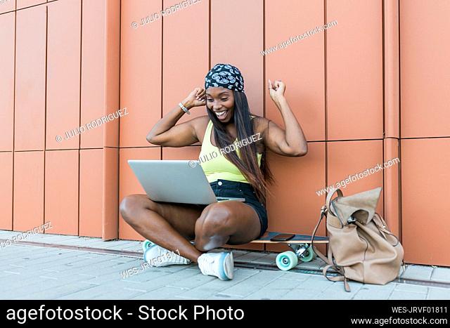 Happy woman cheering while sitting cross-legged on skateboard in front of wall