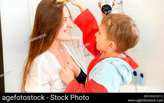 Funny smiling mother and little son drying and combing hair in bathroom at mirror. Concept of child hygiene and health care at home