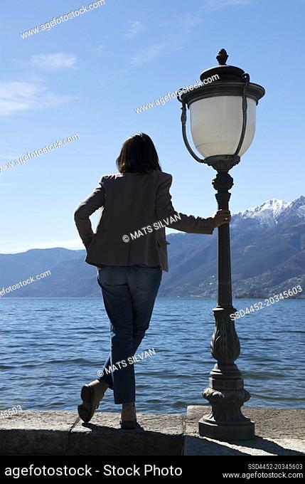 Woman Standing and Leaning on a Street Lamp on Waterfront to an Alpine Lake Maggiore with Mountain in Ascona, Switzerland