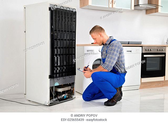 Young Plumber Writing On Clipboard In Front Of Refrigerator Appliance In Kitchen Room