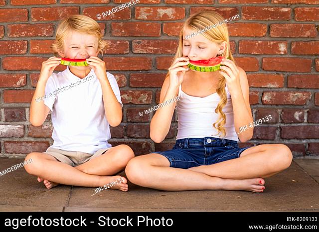 Cute young cuacasian boy and girl eating watermelon against brick wall