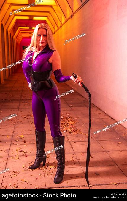 A beautiful 51 year old tall blond dominatrix woman dressed a latex costume, holding a whip in a lighted underpass